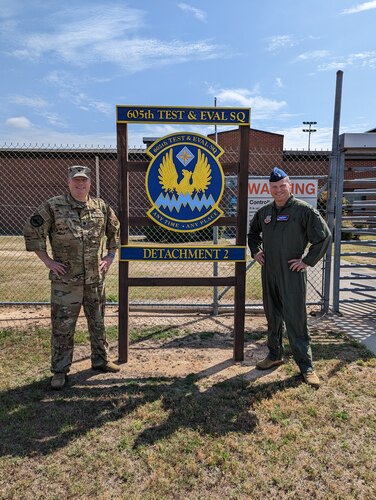 photo of two uniformed U.S. Air Force Airman stand outside gated area next to sign “605th Test and Evaluation Squadron Detachment 2” with unit’s emblem in the center of the sign which states “605th Test & Eval Sq, any time, any place.”