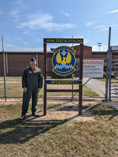 photo of uniformed U.S. Air Force Airman standing outside gated area next to sign “605th Test and Evaluation Squadron Detachment 2” with unit’s emblem in the center of the sign which states “605th Test & Eval Sq, any time, any place.”