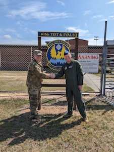 photo of two uniformed U.S. Air Force Airman shaking hands outside gated area next to sign “605th Test and Evaluation Squadron Detachment 2” with unit’s emblem in the center of the sign which states “605th Test & Eval Sq, any time, any place.”