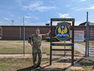 photo of uniformed U.S. Air Force Airman standing outside gated area next to sign “605th Test and Evaluation Squadron Detachment 2” with unit’s emblem in the center of the sign which states “605th Test & Eval Sq, any time, any place.”