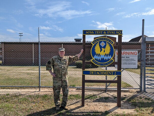 photo of uniformed U.S. Air Force Airman standing outside gated area next to sign “605th Test and Evaluation Squadron Detachment 2” with unit’s emblem in the center of the sign which states “605th Test & Eval Sq, any time, any place.”