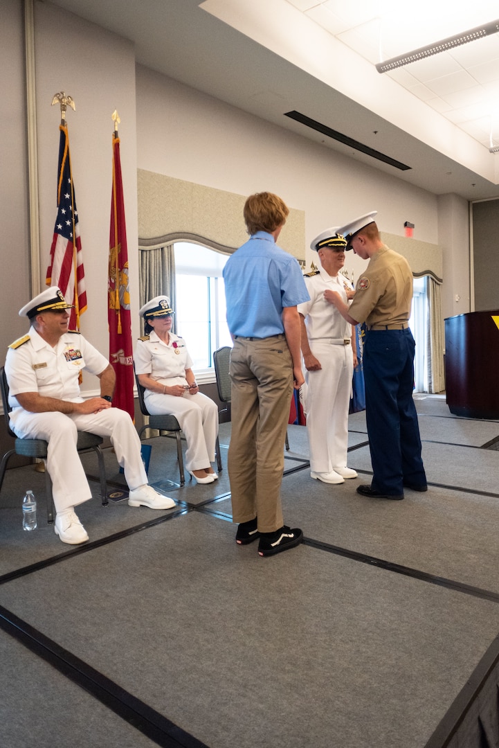 Sean Barbabella, a member of Navy Junior Reserve Officers Training Corps, pins the Command Ashore Insignia upon his father, Capt. Sean Barbabella, during the Naval Health Clinic Cherry Point change of command ceremony May 31, 2023 while Rear Adm. Matthew Case, chief of the Medical Service Corps and commander of Naval Medical Forces Atlantic and the ceremony’s officiant, left, Capt. Elizabeth Adriano, second from left and Matthew Barbabella look on.