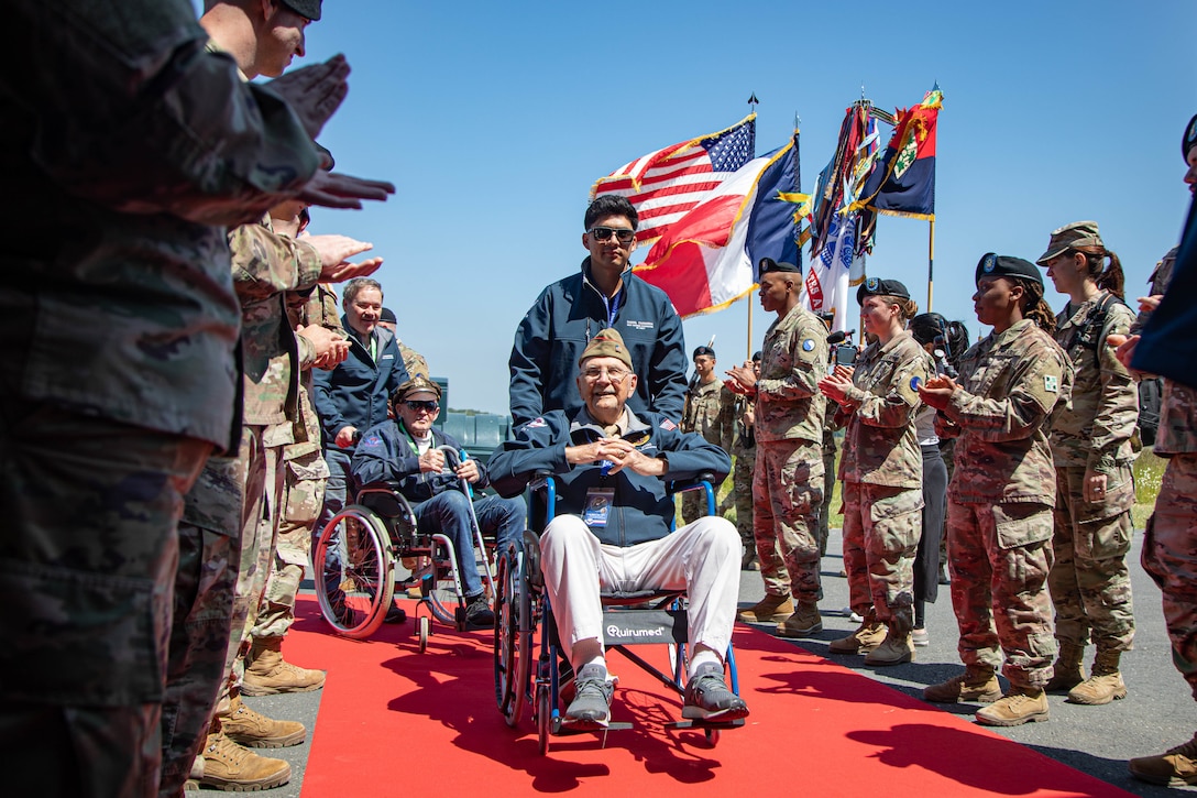 Two men in wheelchairs are escorted on a red carpet through a line of clapping service members.