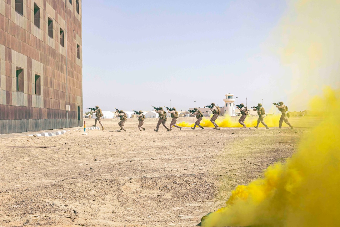 Service members move in a line toward a building as yellow smoke surrounds them.