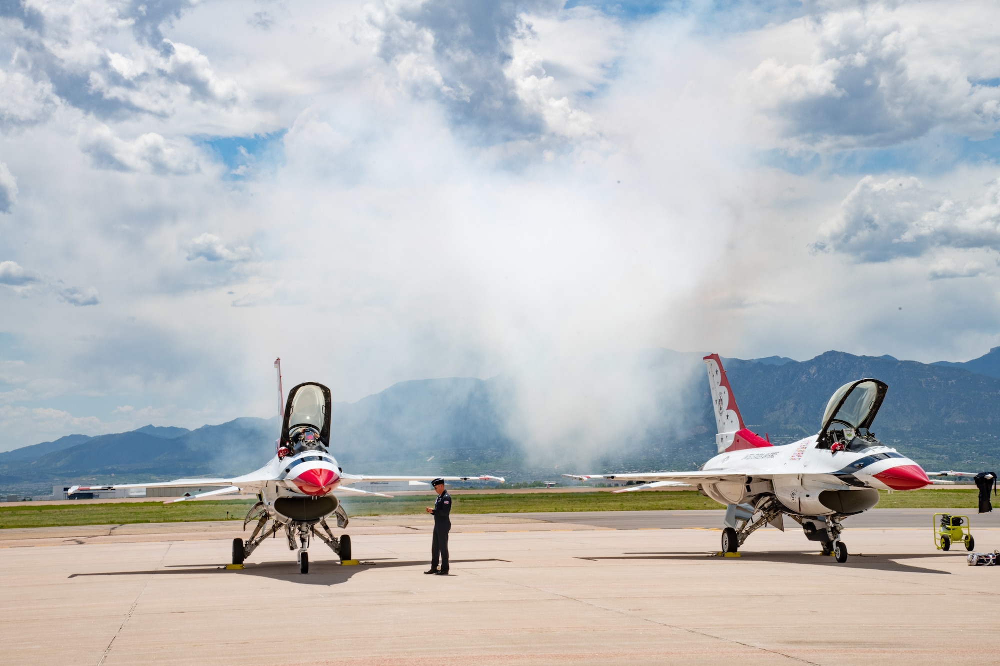Two military aircraft, colored red, white and blue, sit on a tarmac on a sunny day in front of a mountain.