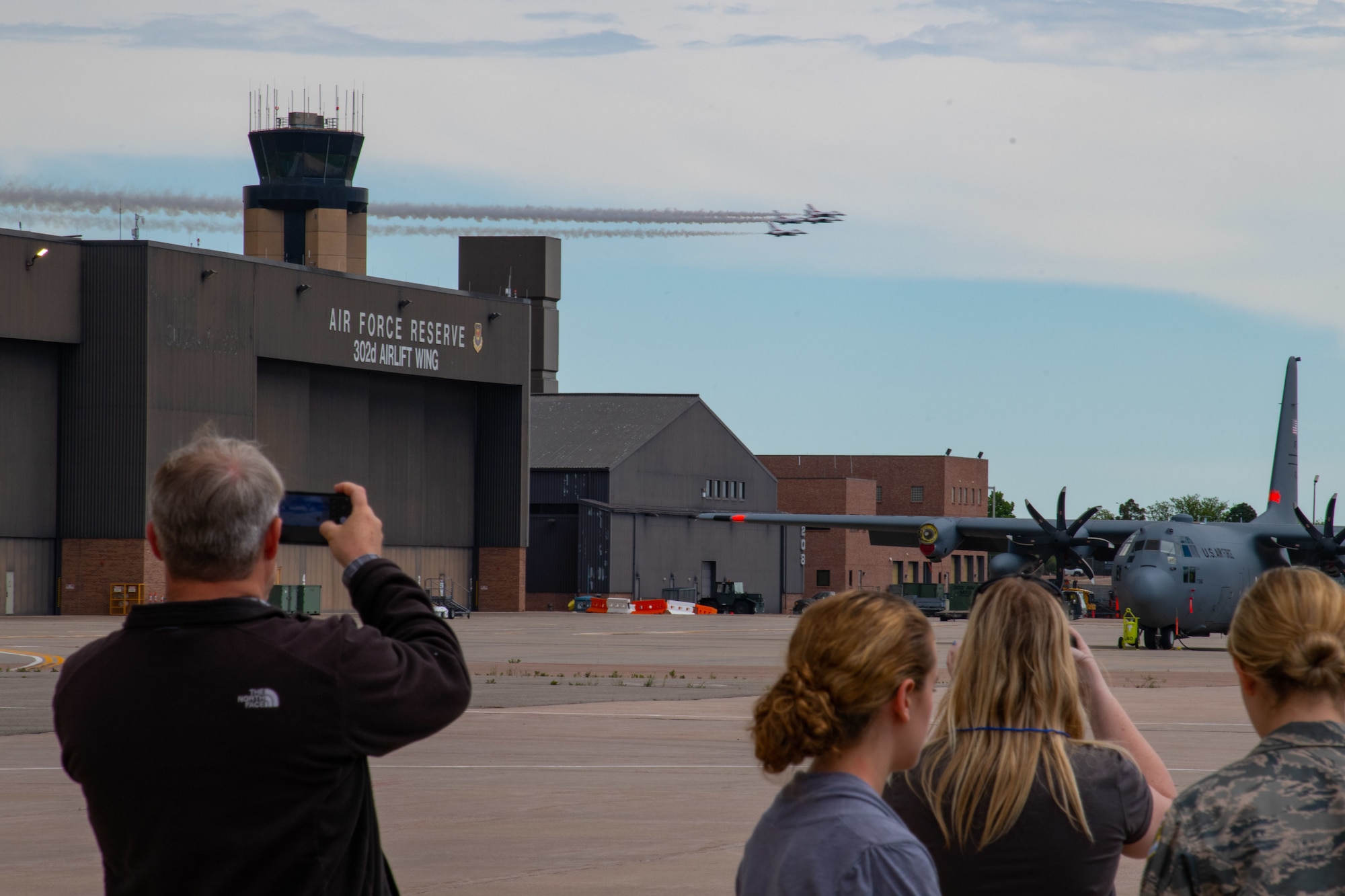 Onlookers stand on flightline and take photos of four military fighter jets flying over a building