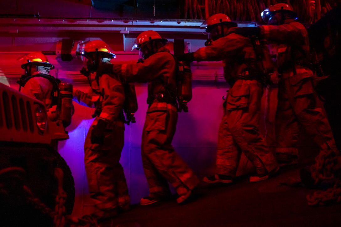 Sailors in firefighting gear walk in a line in a dark area.
