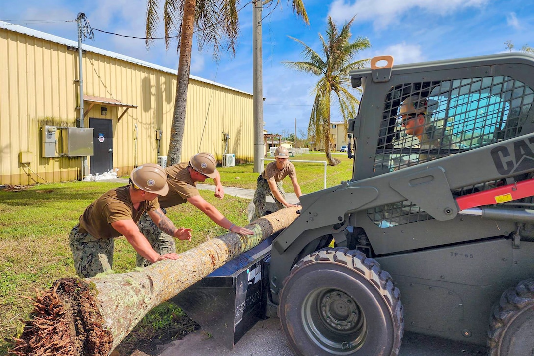 Navy Seabees put a fallen tree onto a skid steer in front of a building.