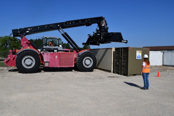 A woman watches a heavy equipment operator lift a large shipping container with a tractor.