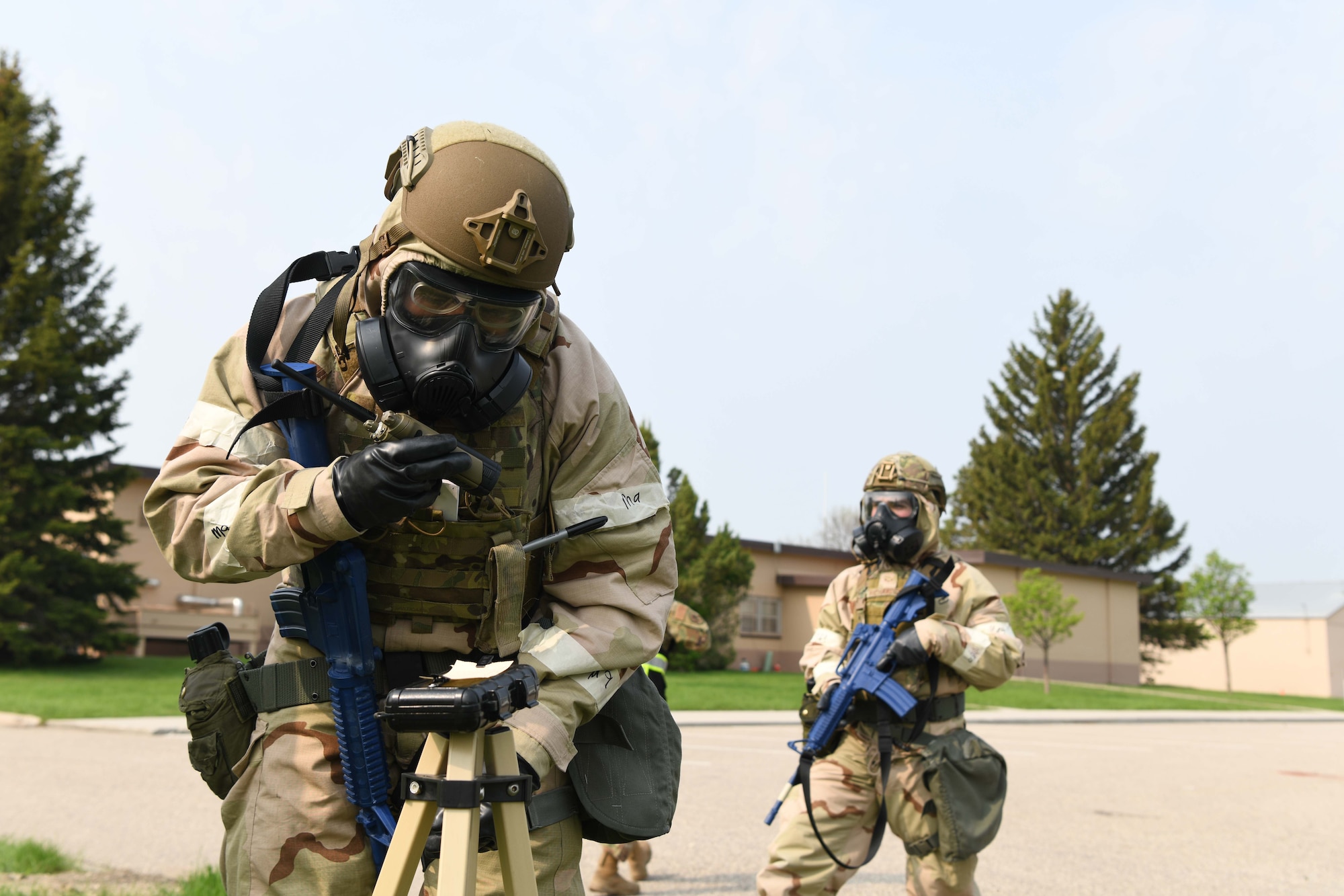 One airman in CBRN gear checks paper for chemical contamination while another airman in CBRN gear holds a blue training rifle.