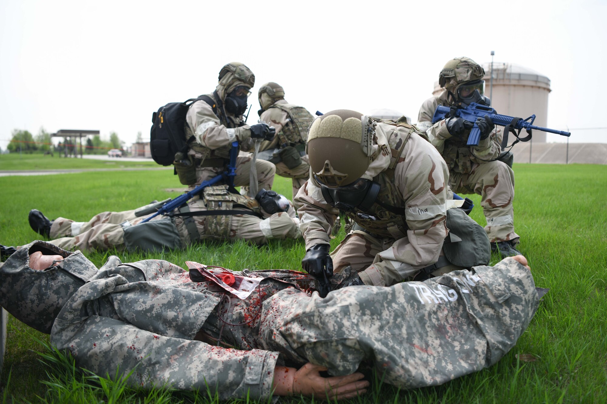 Two airmen in CBRN gear administer medical aid to two mannequins on green grass.