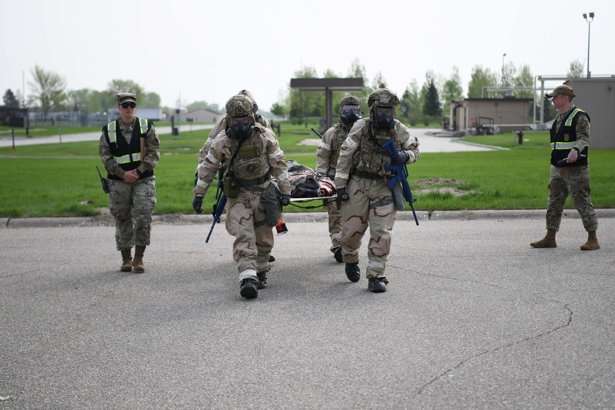 Four airmen in CBRN gear carry a litter while walking on grey pavement.