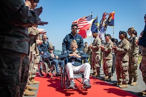 Two men in wheelchairs are escorted on a red carpet through a line of clapping service members.