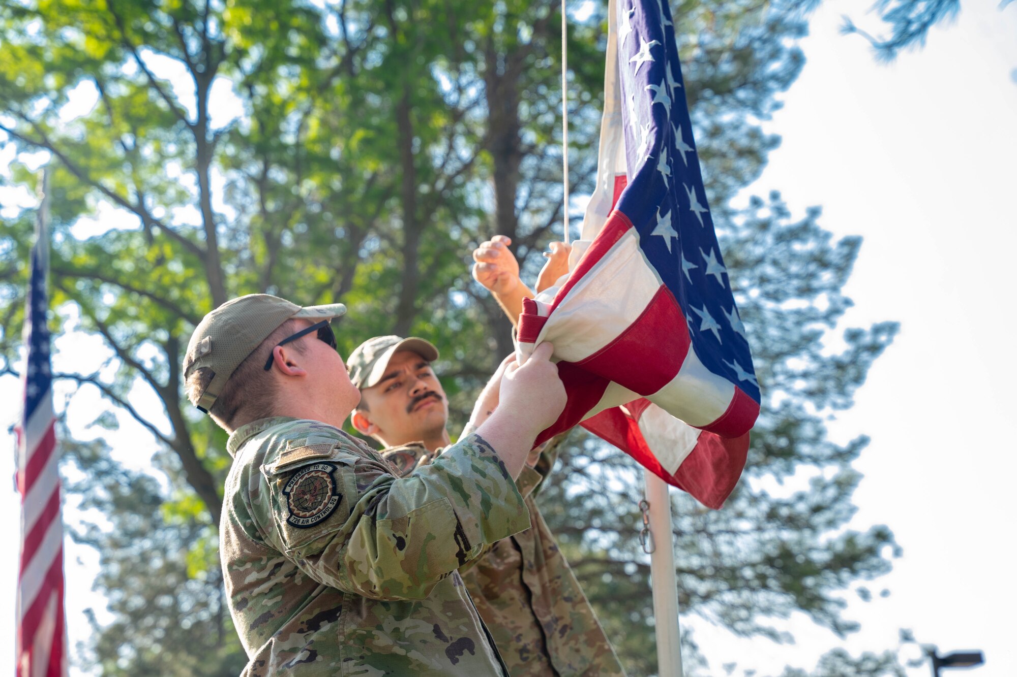 Senior Airman Chase Hanes, left, 726th Air Control Squadron, and Senior Airman Jonathon Vowels, 366th Security Forces Squadron, Mountain Home Air Force Base, hoist up the flag at the Idaho State Veterans home, Boise, Idaho, May 20, 2023. Airmen volunteered for a car show event at the veteran’s home, assisting senior veterans with voting on their favorite car. (U.S. Air National Guard photo by Senior Airman Natalie Filzen)