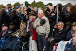 A group of World War II veterans and others, some seated, others standing, salute and pay respects during a ceremony.