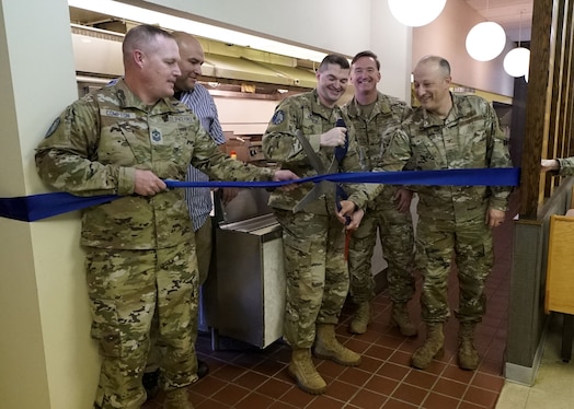  Four men in green military uniforms and a man in a blue shirt cut a large blue ribbon with large silver scissors.