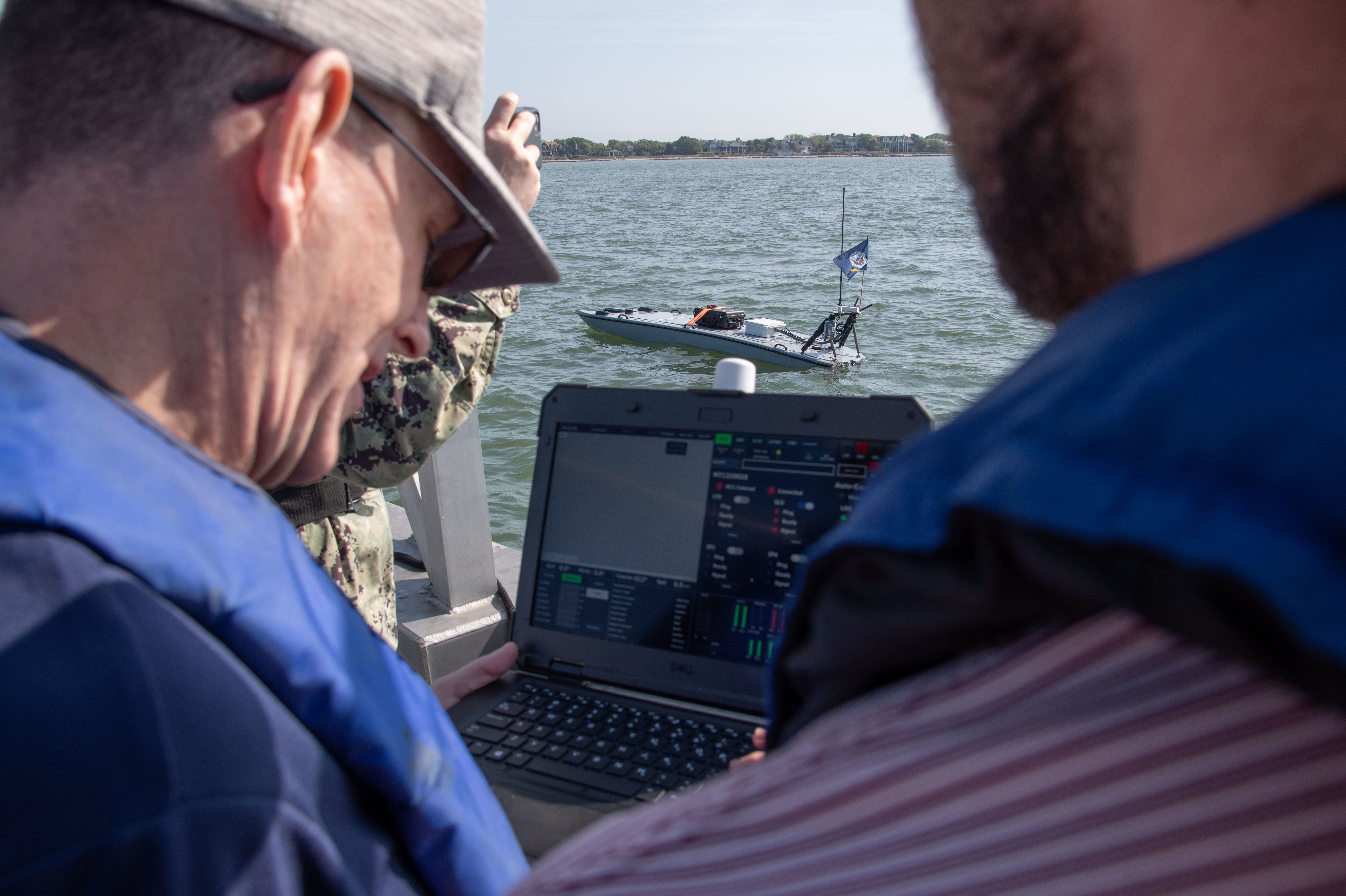 Mr Reddy on the left observes a laptop interface. In the background sits an unmanned surface vessel in the water.