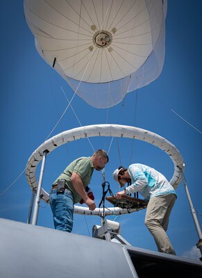 Two men working on radio equipment to a ring a the bottom of a balloon.