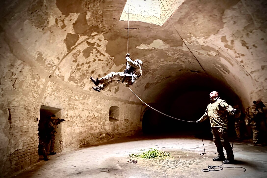 A soldier rappels into a stone fort while another holds the rope.