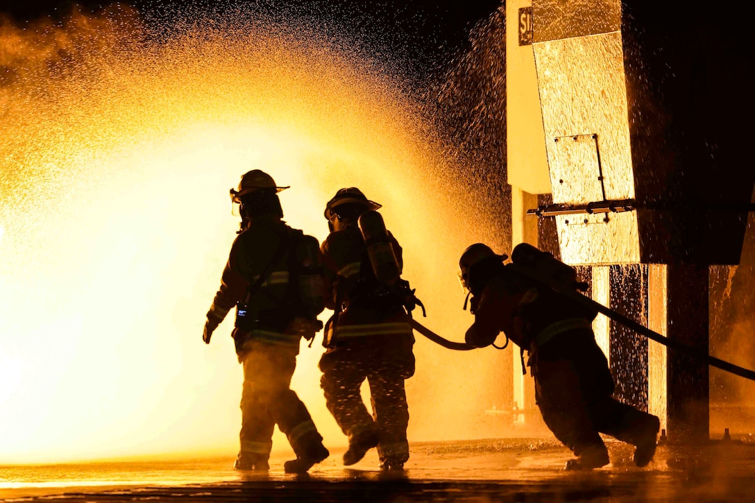 Three Marines shown in silhouette wearing fire protection equipment use a hose to spray water.