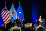 Two men sit on a stage with several flags behind them.