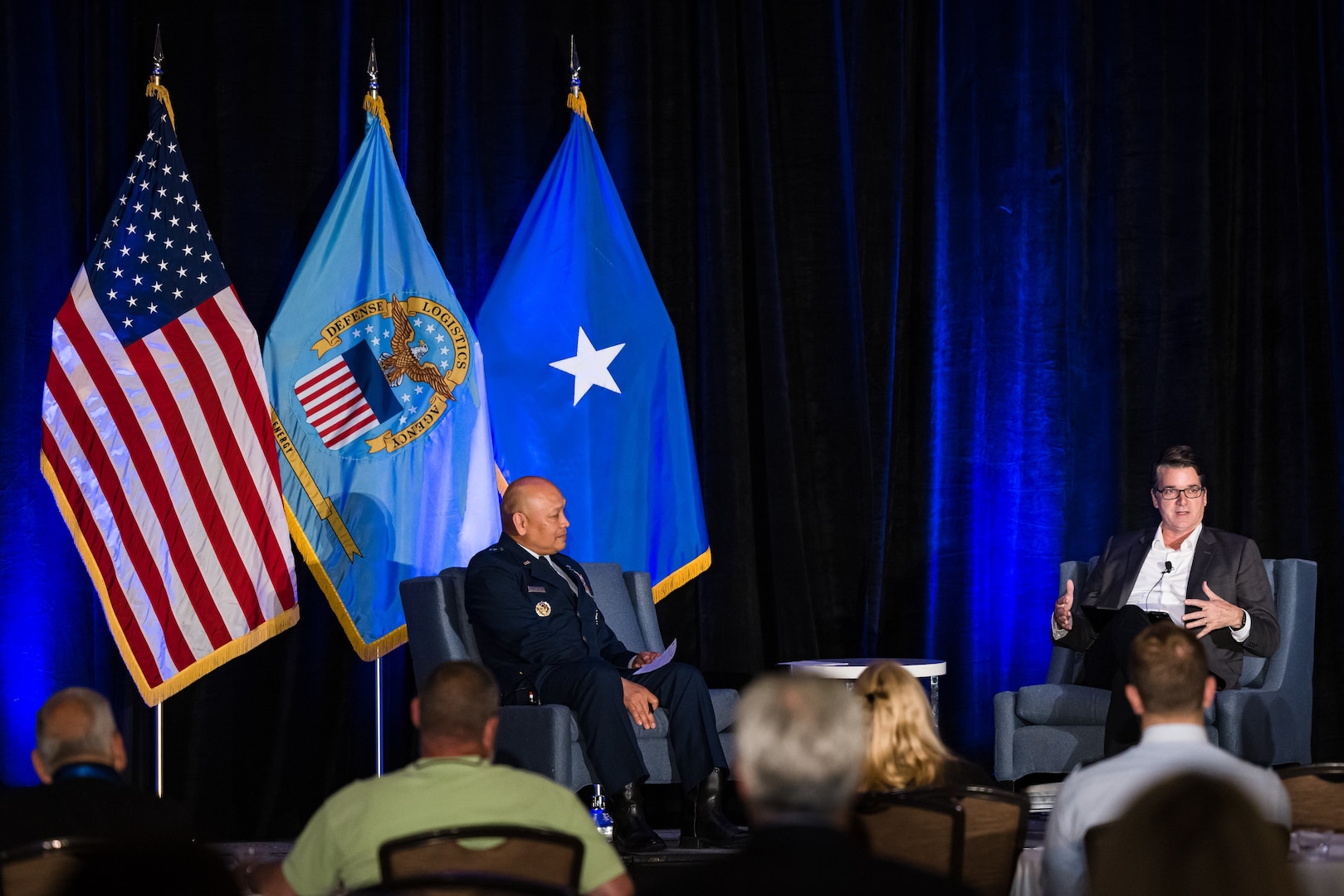 Two men sit on a stage with several flags behind them.