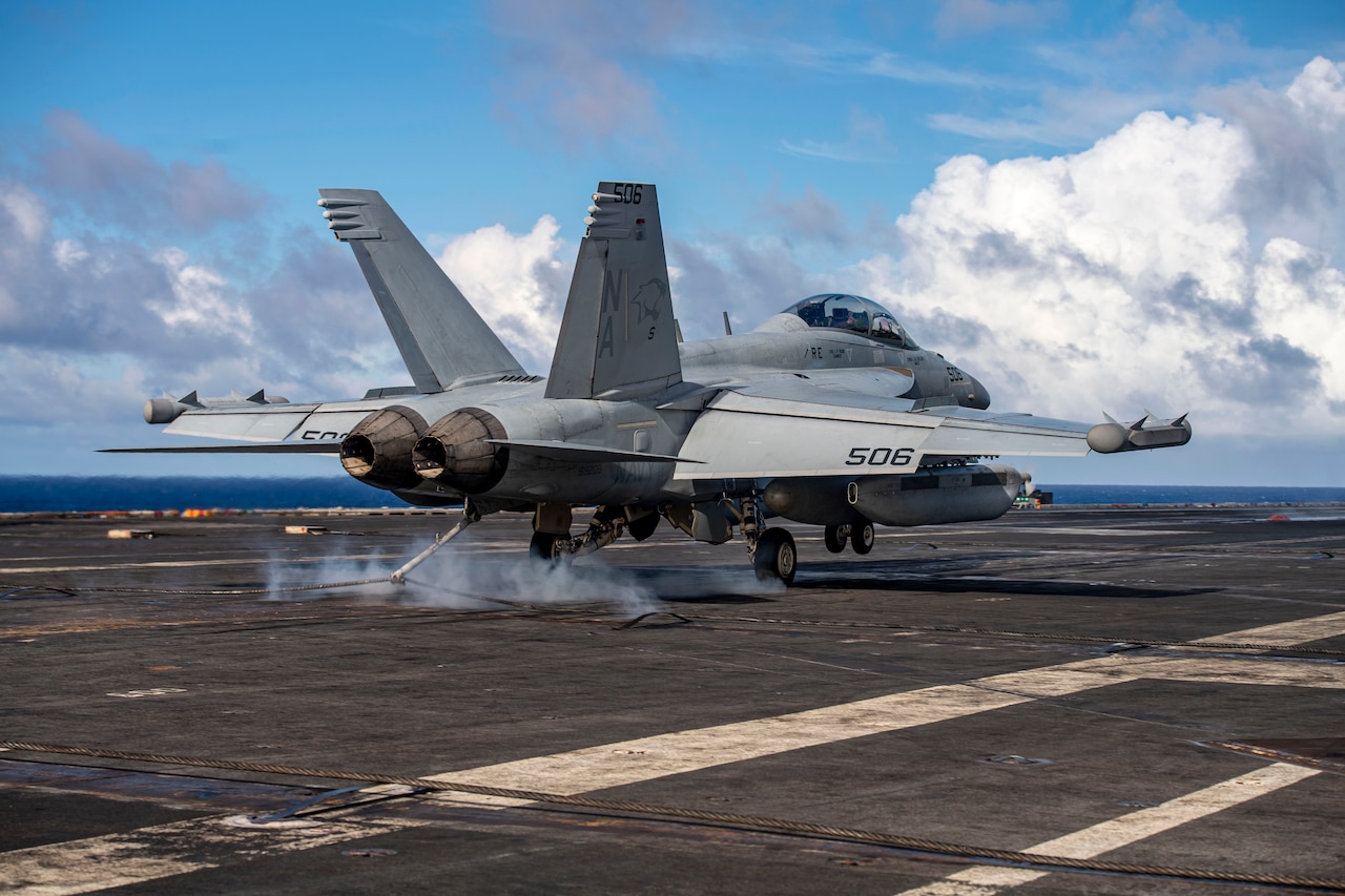 Side-rear view of an aircraft landing on the deck of a ship at sea.