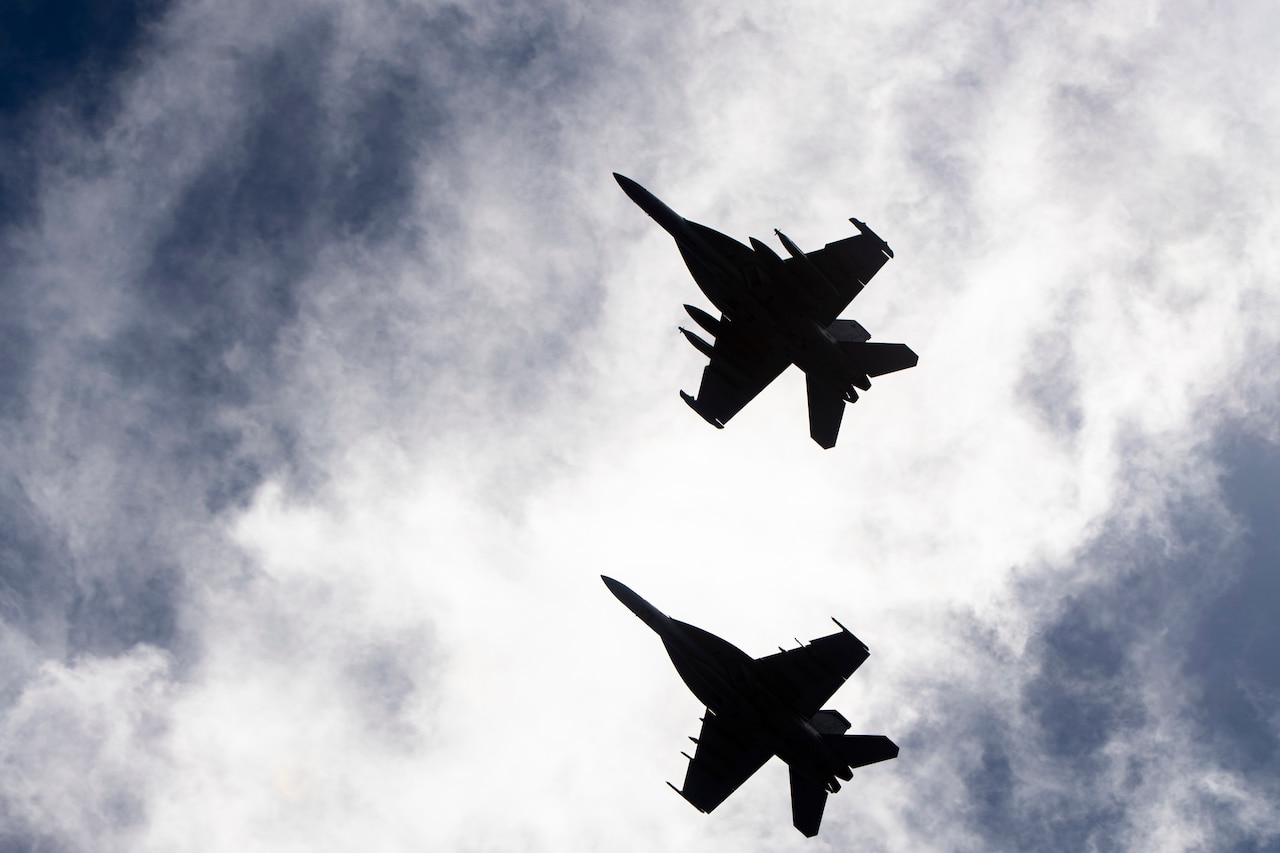 Two aircraft shown in silhouette fly next to each other in cloudy sky.