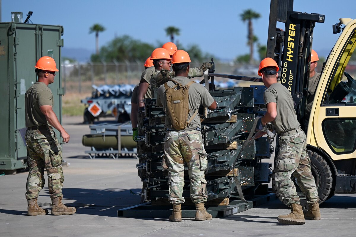 A photo of a group of people in military uniforms helping guide a forklift driver to lift part of a munitions assembly conveyor.