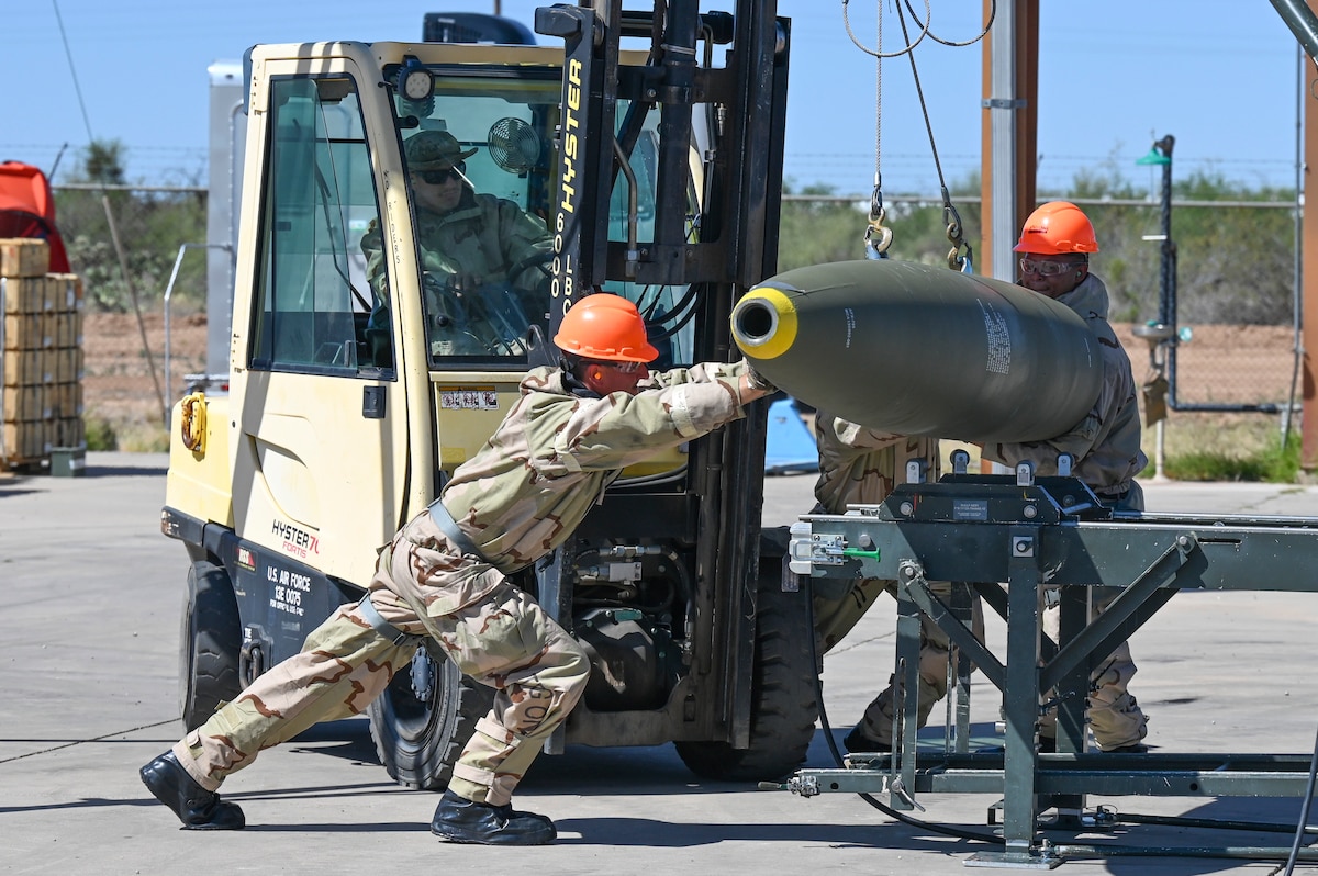 A person in military hazardous material protective gear pushes a bomb suspended from a fork lift onto a munitions assembly conveyor belt.