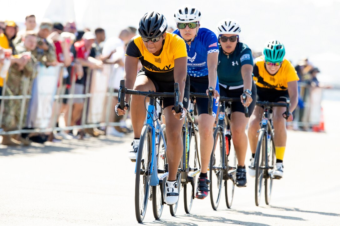 Four service members ride bikes in formation as spectators observe from behind a barrier.