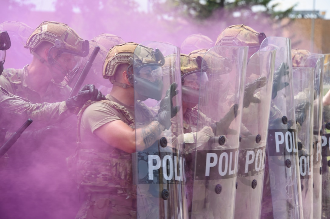 National Guardsmen wearing protective gear form a shield wall surrounded by purple smoke.