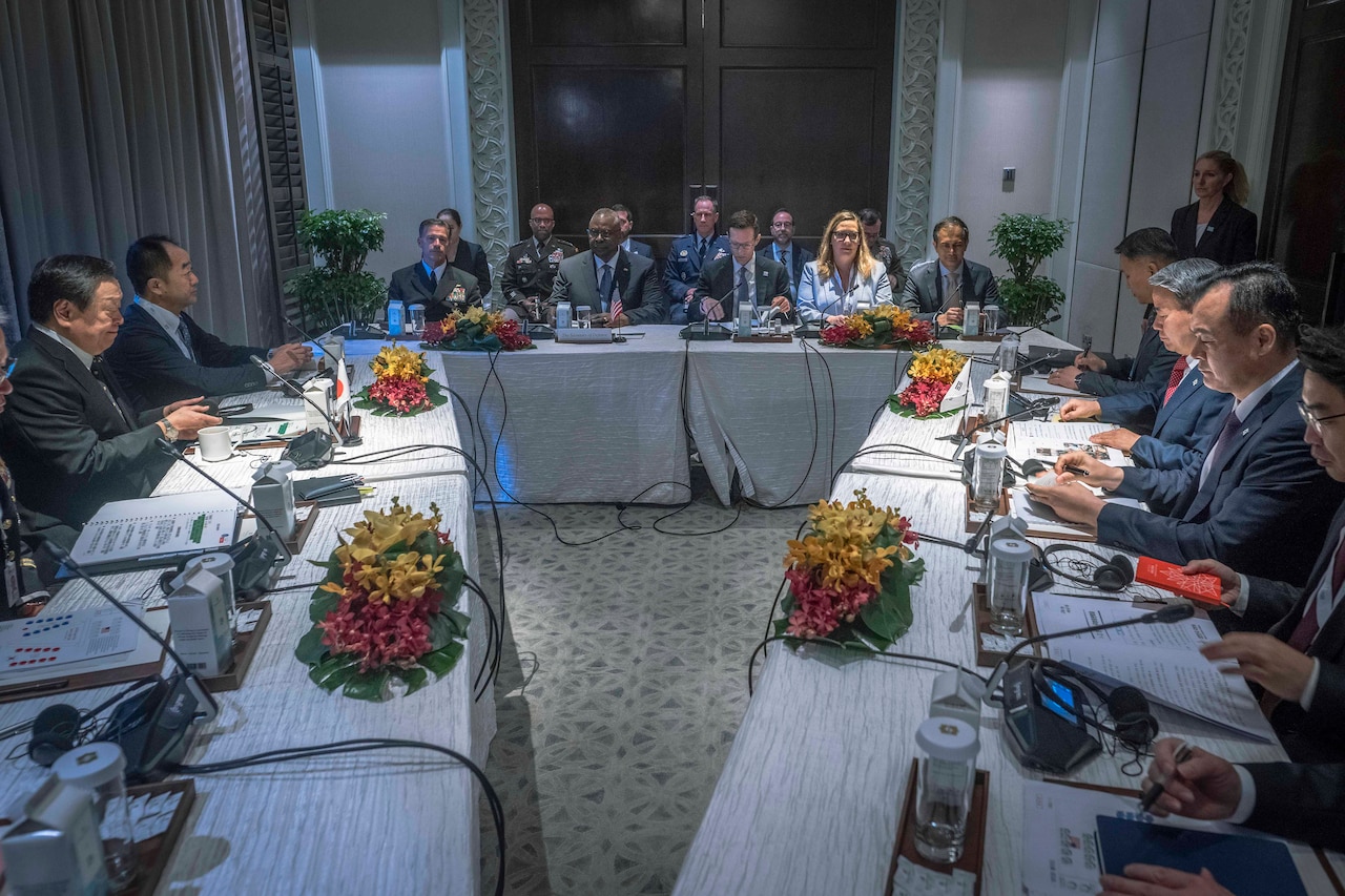 People in business attire and military uniforms sit at tables arranged in a triangle.