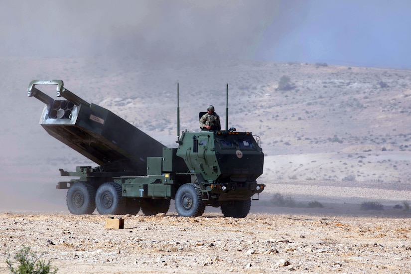 A service member looks out from the top of a military vehicle which sits in a barren, desert landscape.