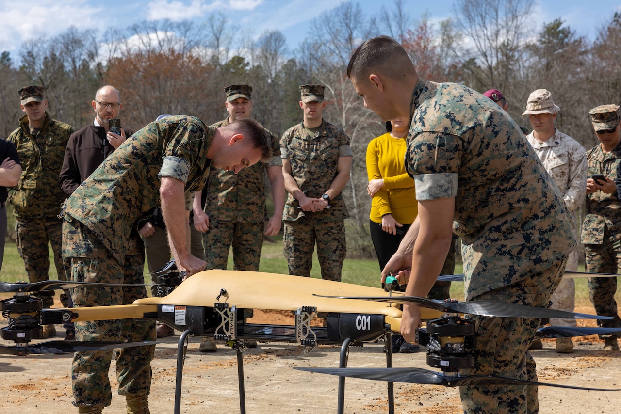 Two uniformed service members handle a drone in front of a group of uniformed service members and civilians.