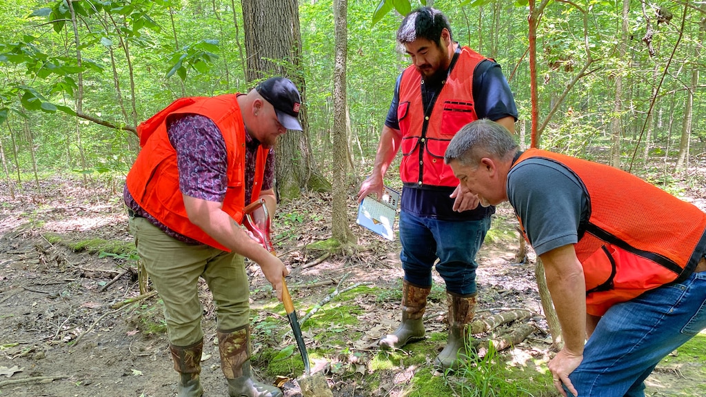 Three men gather in a circle around a patch of soil. One has a shovel, another has a Munsell Book, and the other is observing.