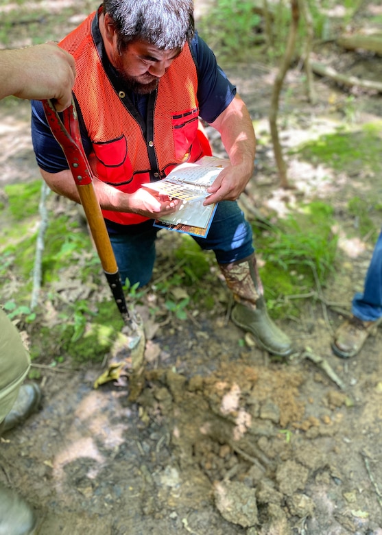 A man stands on a soil bed comparing colors of soil to a chart.