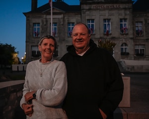 A French lady in a blue sweater stands next to her husband in a black sweatshirt outside the town hall in Sainte Mere Eglise, France.
