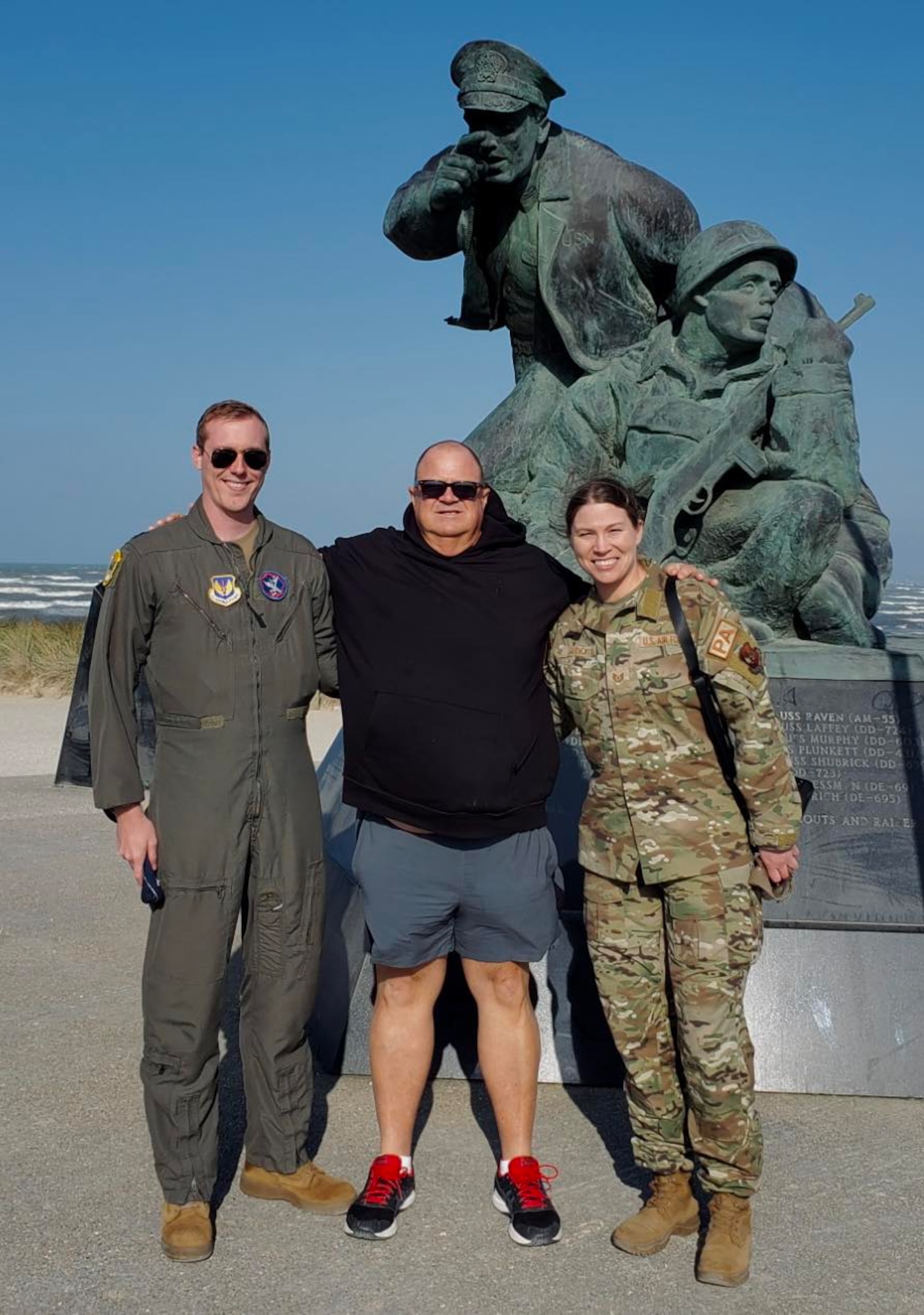 Two Airmen, one in a flight suit and one in OCPs, stand on either side of a man in a black sweatshirt next to a monument a Utah Beach, Normandy, France.