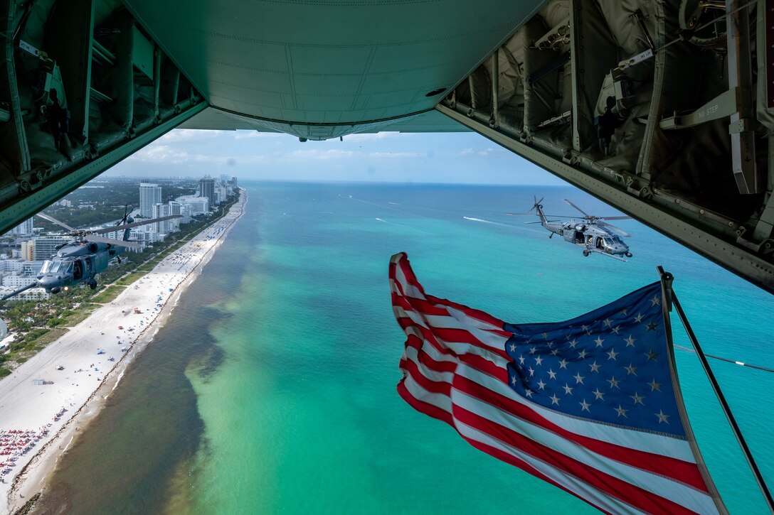 Two helicopters fly behind a large aircraft carrying the American flag above a beach.