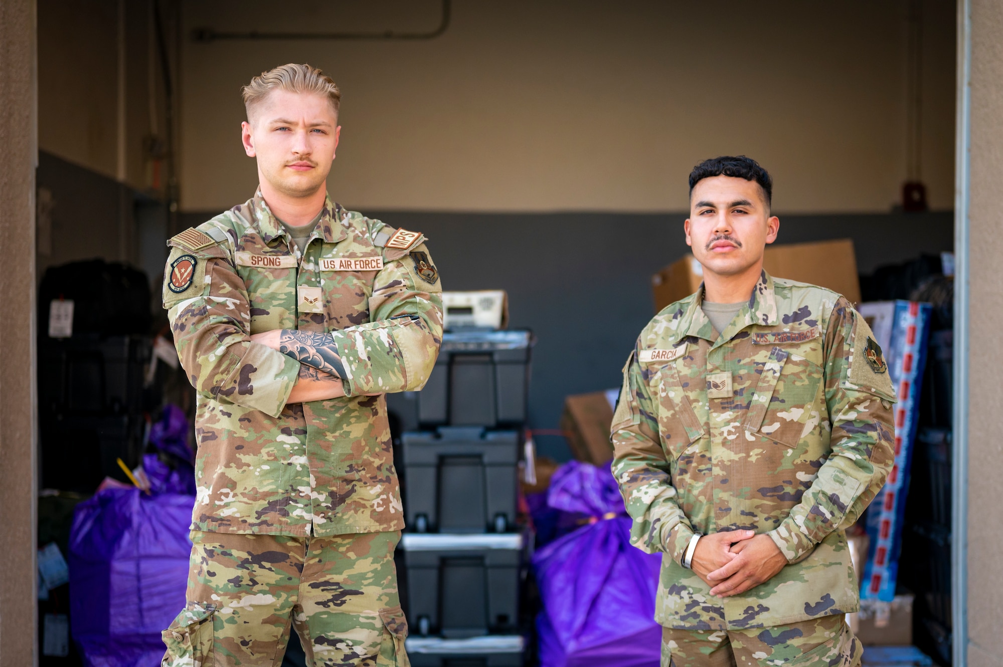 Two military members stand outside in front of a wall of packages and mail.