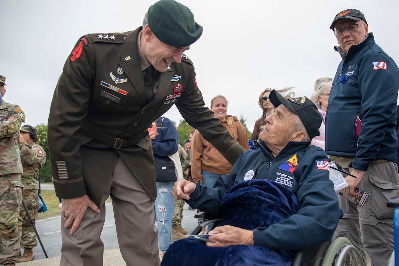 A uniformed soldier greets a seated WWII veteran during an outdoor D-Day ceremony.