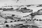 Ships and military personnel land on a beach in a black and white photo.