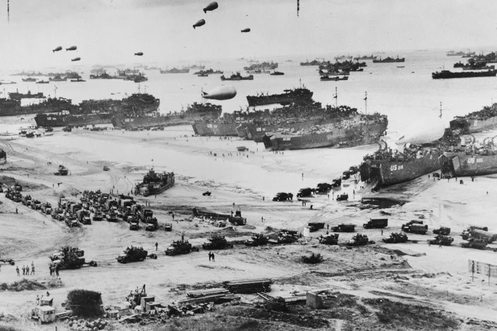 Ships and military personnel land on a beach in a black and white photo.