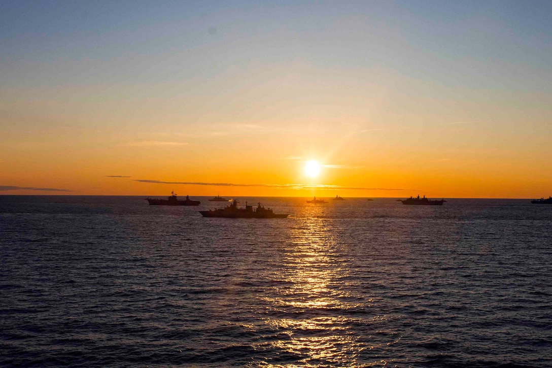 Ships travel through a body of water in formation as the sun shines near the horizon.