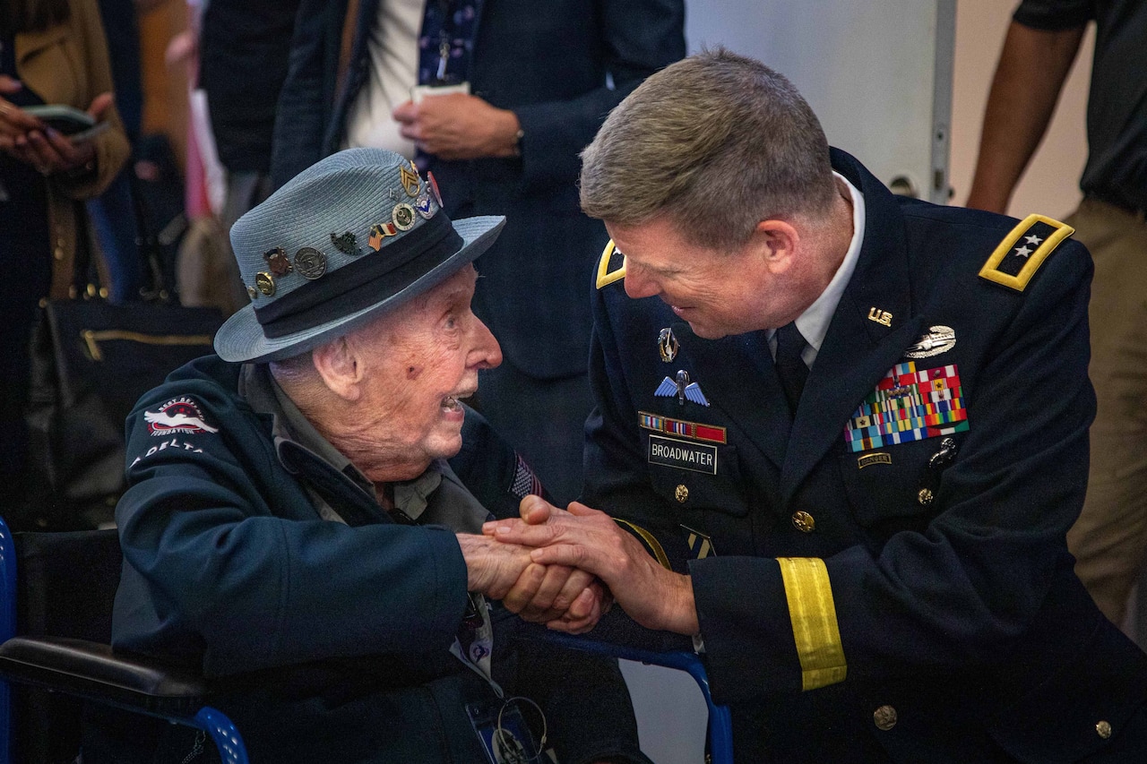 A uniformed service member shakes hands with a seated World War II veteran during an outdoor ceremony.