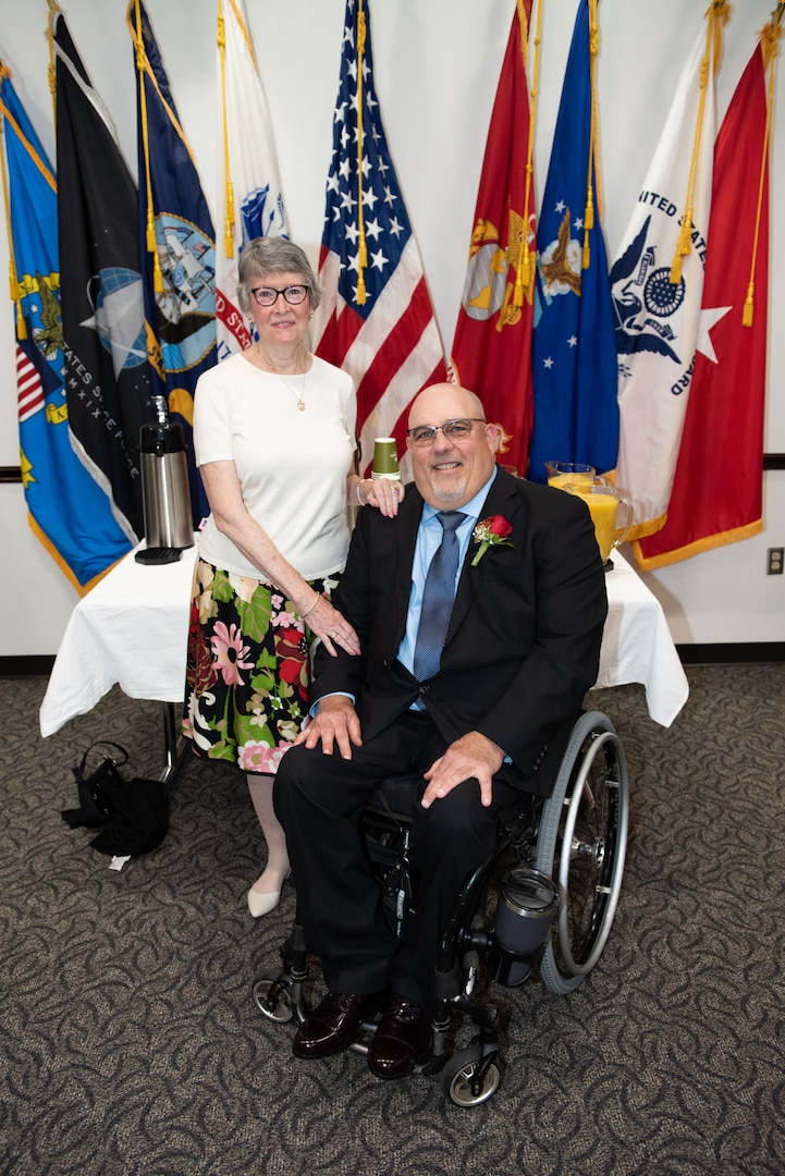 A woman stands by a man in a wheelchair in front of flags.