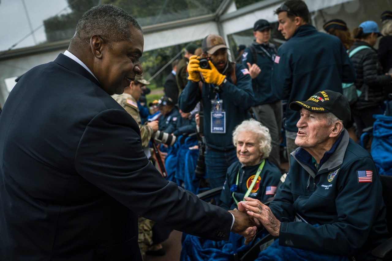 A smiling person in a suit leans forward to shake hands with a seated World War II veteran during a D-Day ceremony.