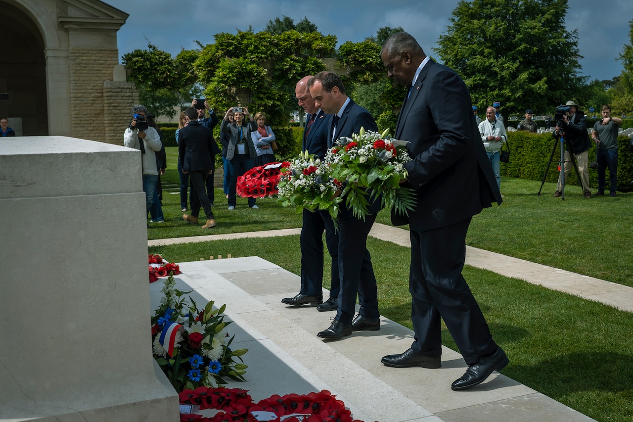 Three people in suits lay wreaths during a D-Day remembrance ceremony.