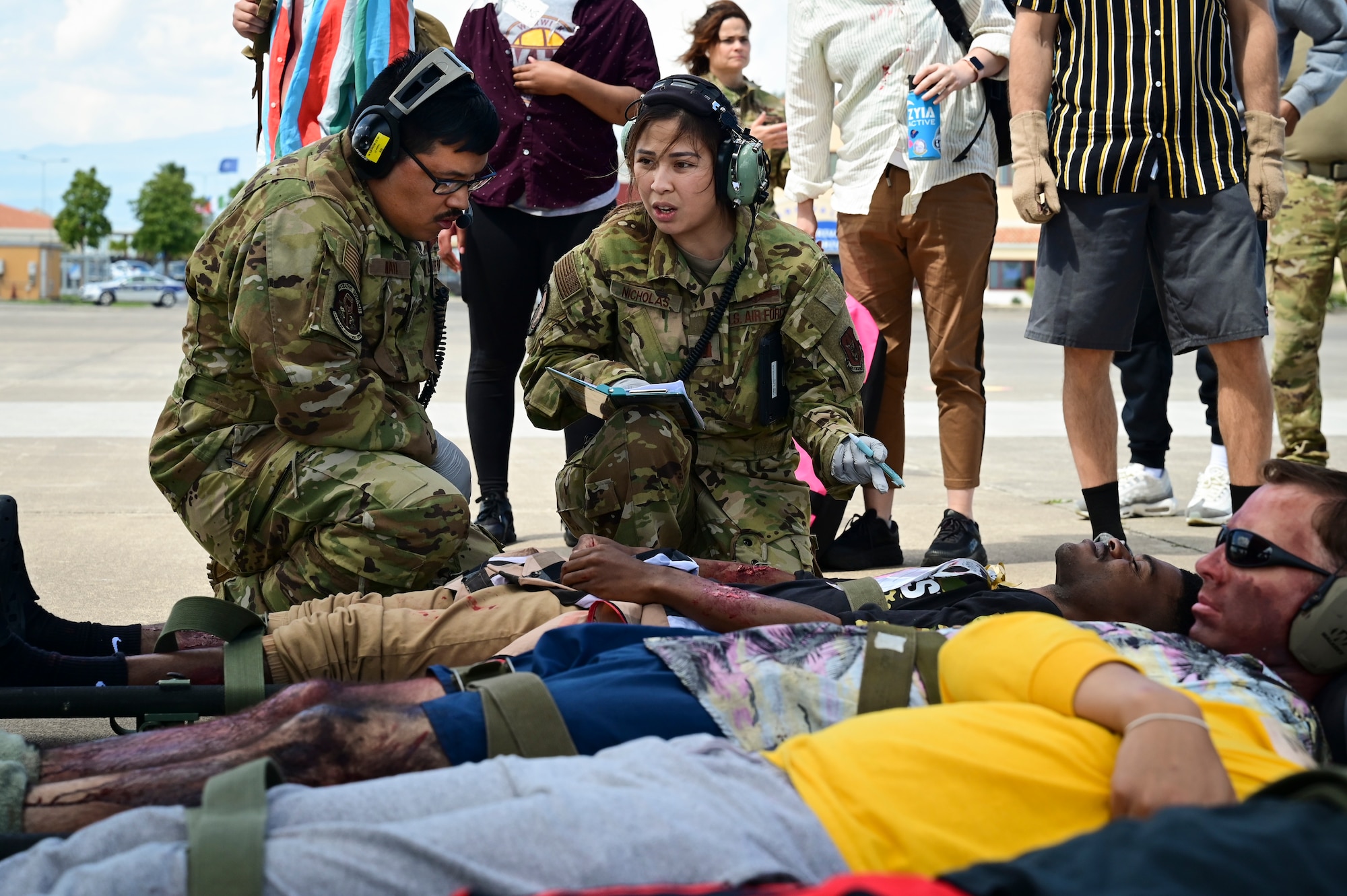 Two Airmen kneel by three stretchers as volunteer patients pretend to be injured for assessment as people watch from behind.
